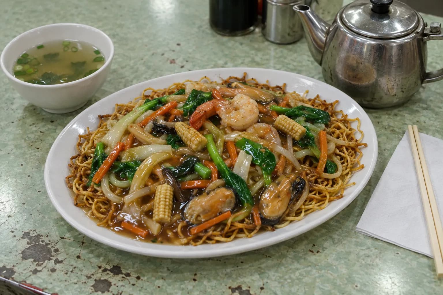 A plate of pan-fried noodles with vegetables and a small bowl of soup on a formica table