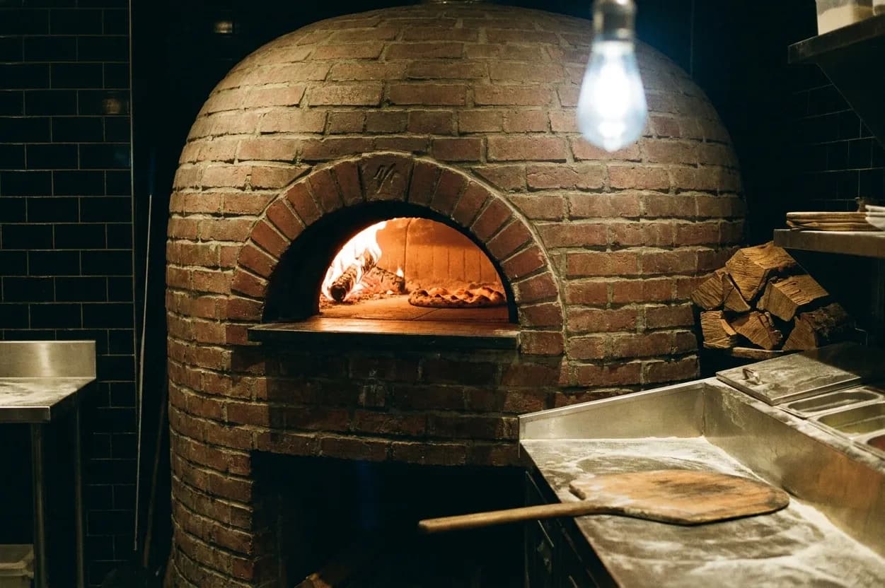 The glowing arched mouth of a brick wood-fired pizza oven in a cramped pizzeria kitchen, with a wooden peel resting on a floured stainless bench
