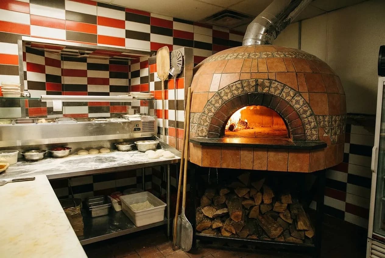 A small pizzeria kitchen with a tiled domed oven glowing orange behind a flour-dusted stainless prep bench