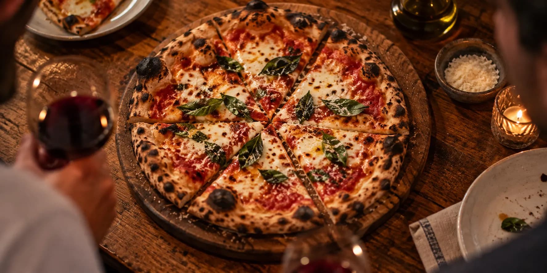 A thin-crust pizza on a wooden board with charred edges, torn basil and a light dusting of grated cheese, cut into wedges