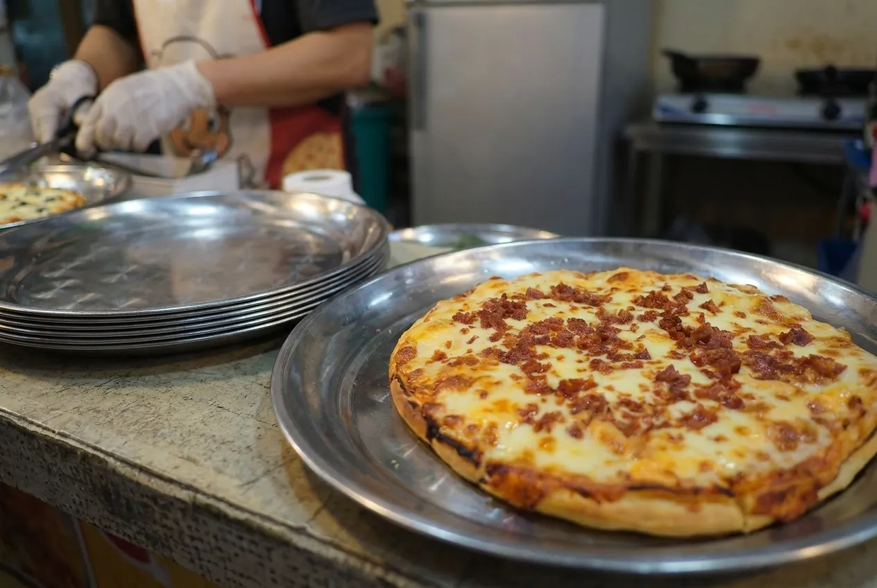 A pizza on a metal tray on a counter beside stacked plates