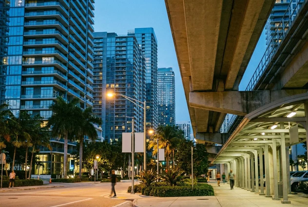 Glass residential towers rising directly from a landscaped street in Brickell at dusk, a covered walkway and the underside of an elevated rail line in frame