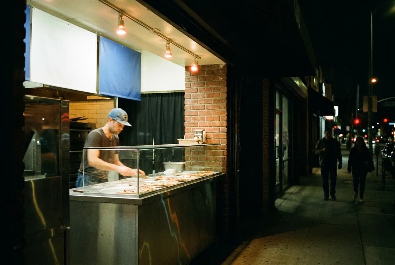 A lit pizza counter seen from a dark pavement late at night
