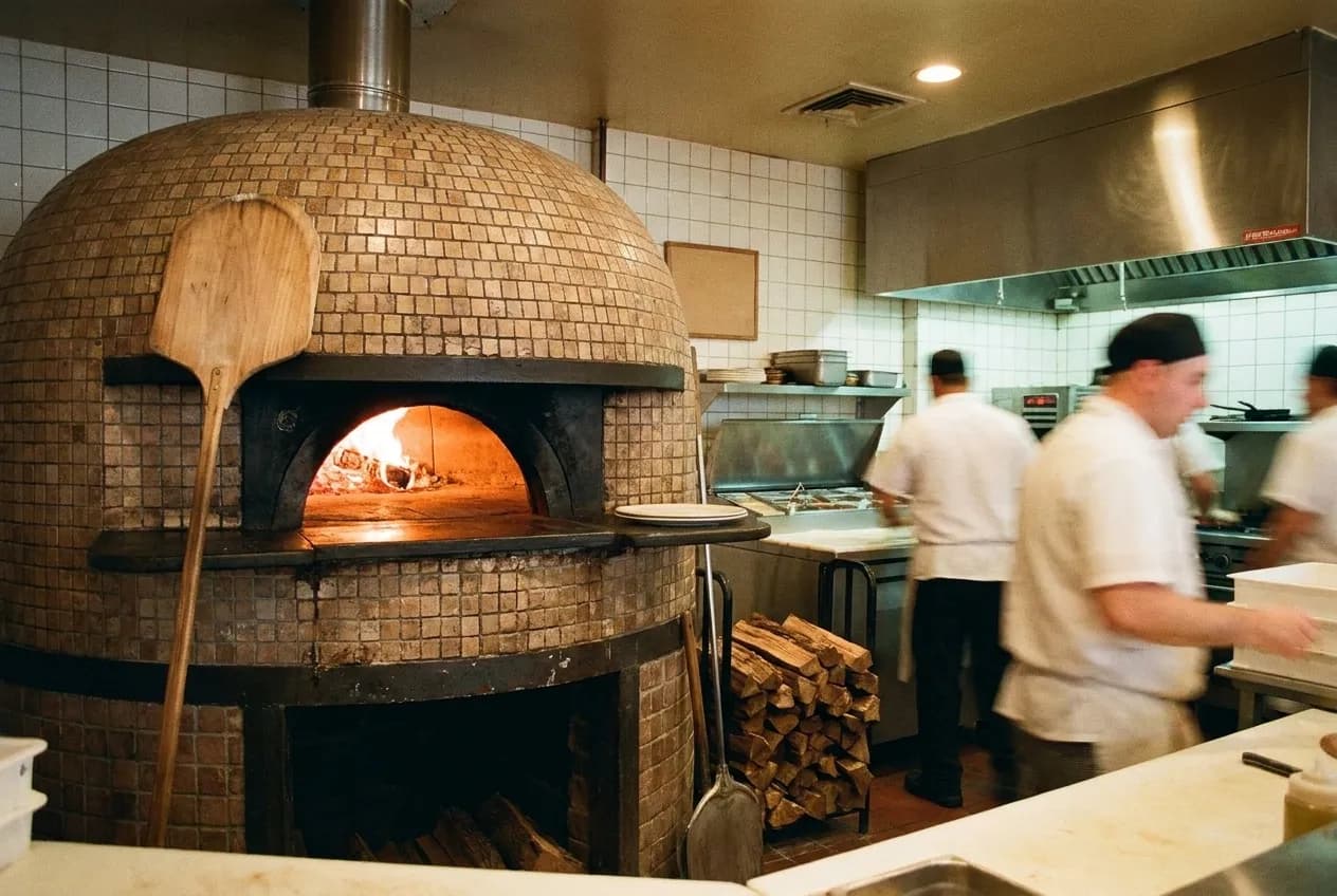 The mouth of a domed wood-fired oven glowing orange inside a small pizzeria, tiled front and a wooden peel resting against the brick