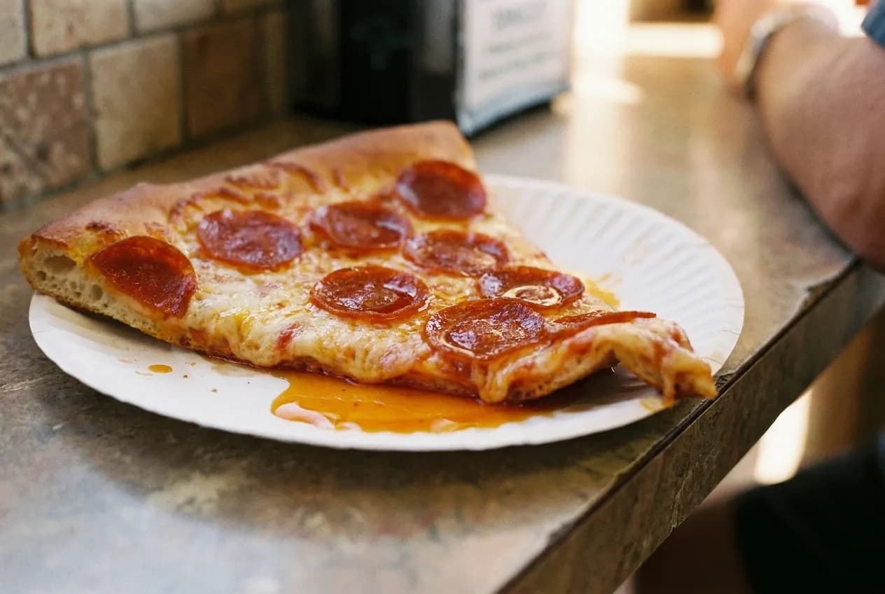 A single wide slice of pizza on a paper plate on a laminate counter, oil pooled in the cheese and the tip beginning to fold