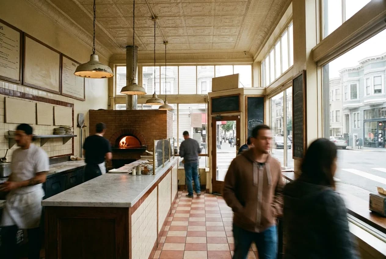 The interior of a small pizzeria with a counter along one side and the mouth of an oven glowing at the back, tiled surfaces and pendant lamps overhead