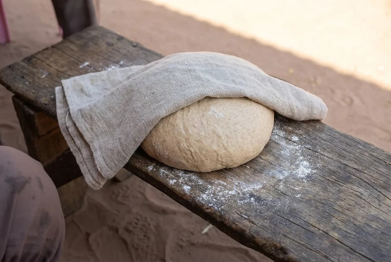 A ball of dough resting under cloth on a floured wooden bench