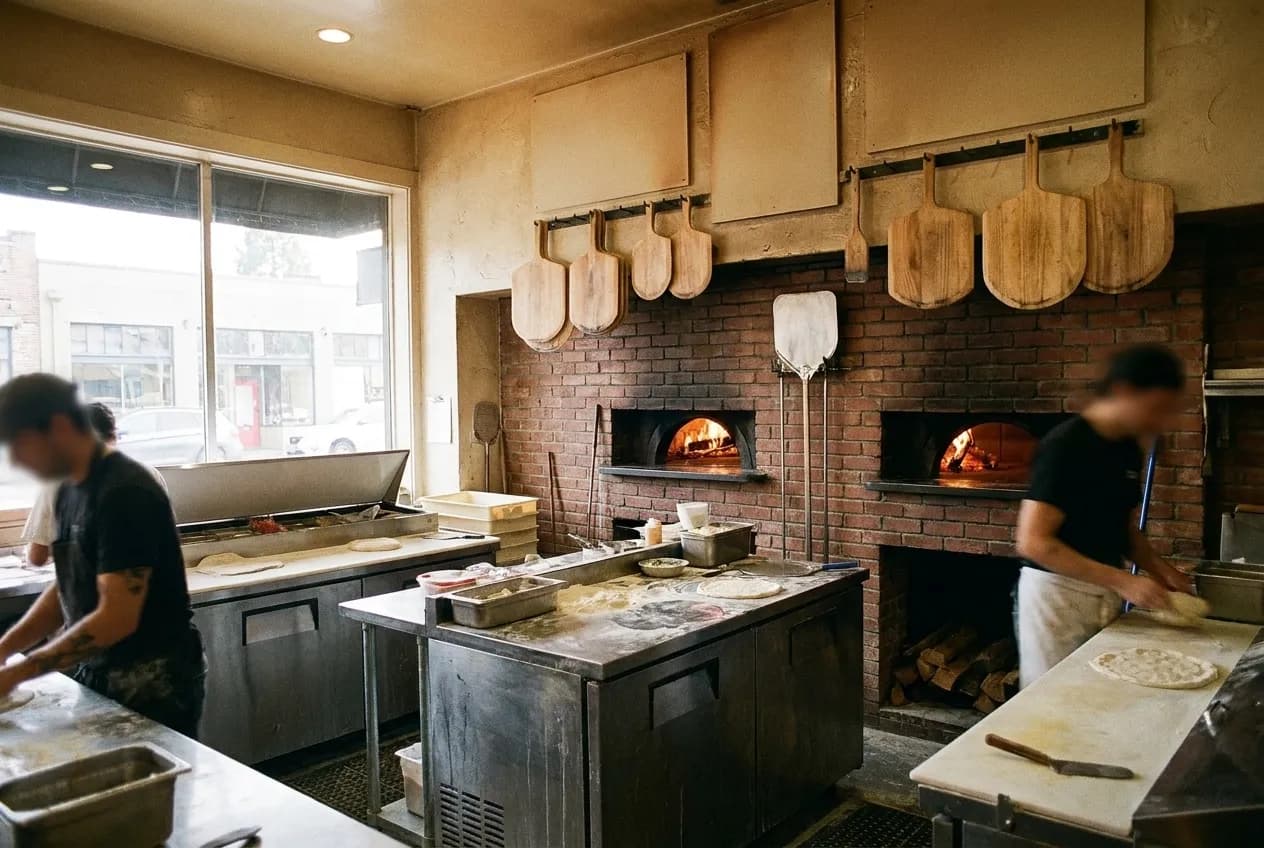 A working pizzeria kitchen with more than one oven mouth visible along the back wall, stainless benches in the foreground and peels hanging within reach