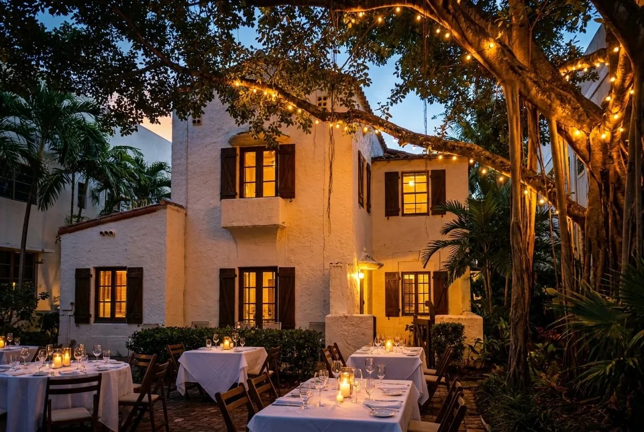 A banyan tree shading a candlelit courtyard patio with string lights, outdoor tables, and a historic stucco facade in Coral Gables at twilight.