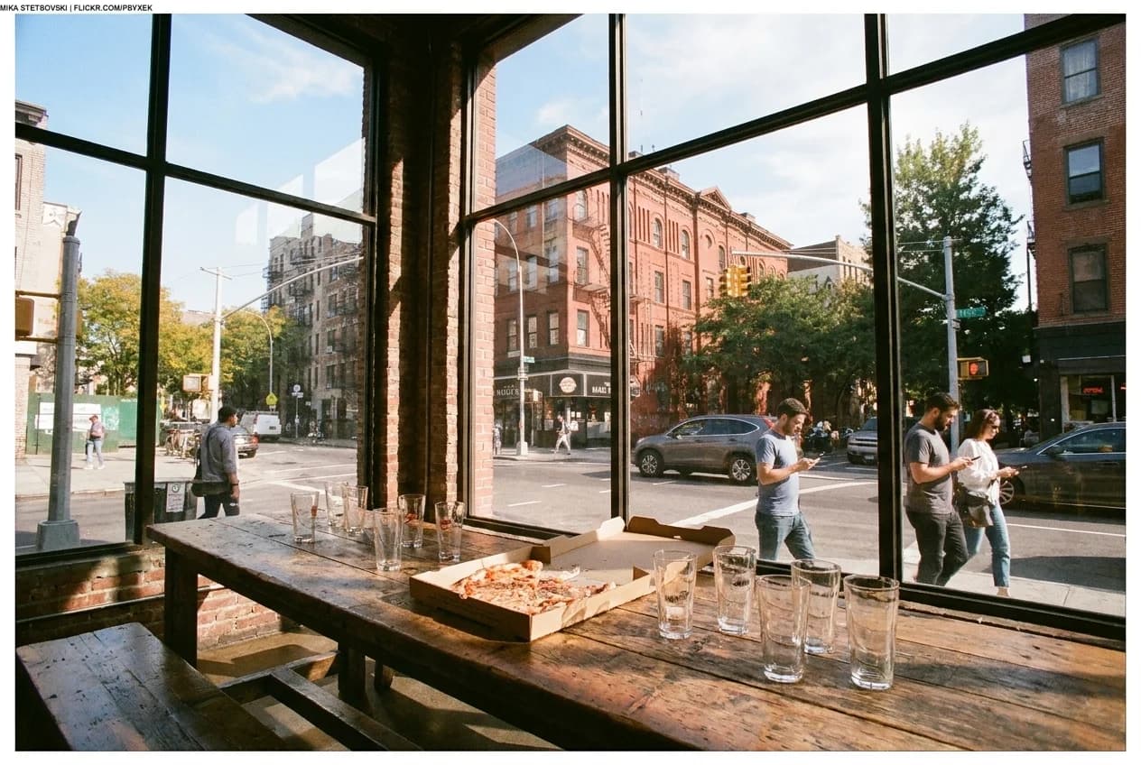 Communal table in Williamsburg taproom with pizza box and pint glasses under natural light