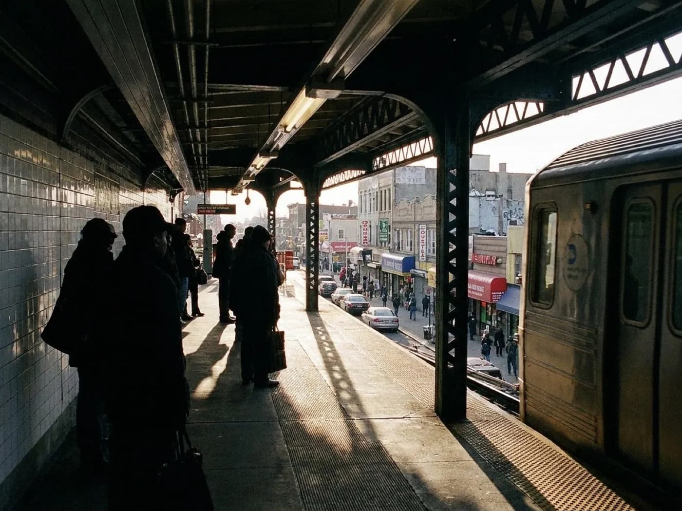 An elevated subway platform above a busy commercial street with shopfronts and awnings below, seen in late afternoon light