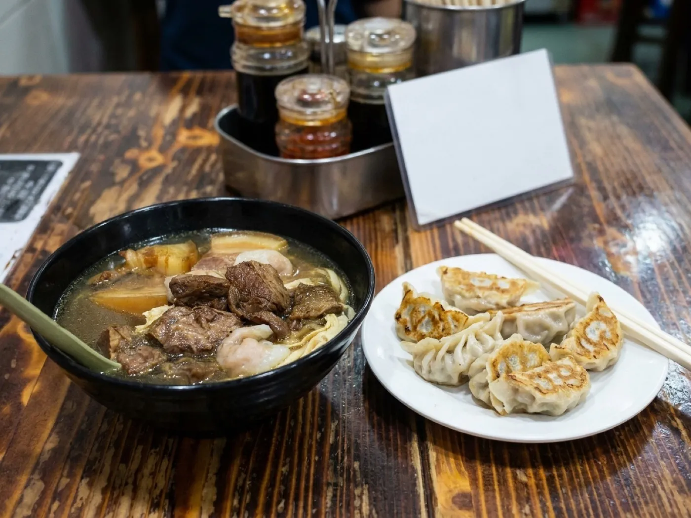 A plate of dumplings and a bowl of noodle soup on a formica table in a small restaurant with a laminated menu propped behind