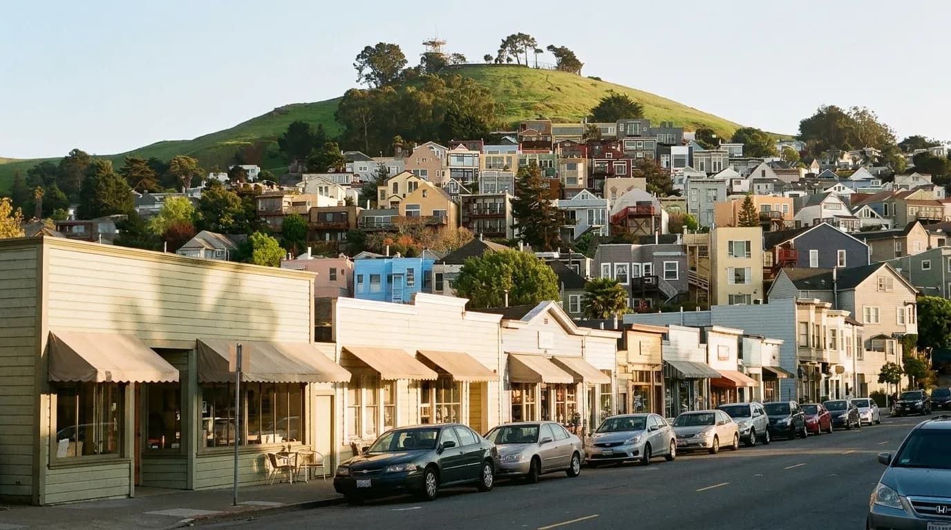 A short neighbourhood shopping street of low painted shopfronts with a steep hill of houses rising behind