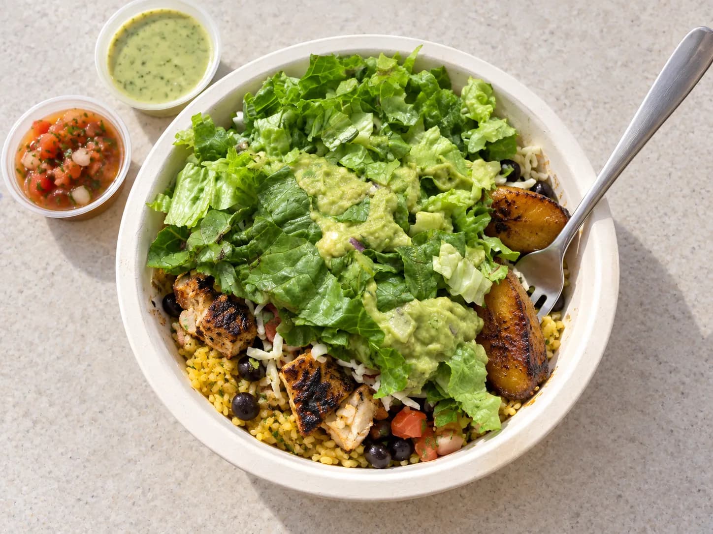 Layered bowl of yellow rice, black beans, citrus-marinated grilled chicken and fried sweet plantains in a plain white takeaway bowl with sauce cups beside it