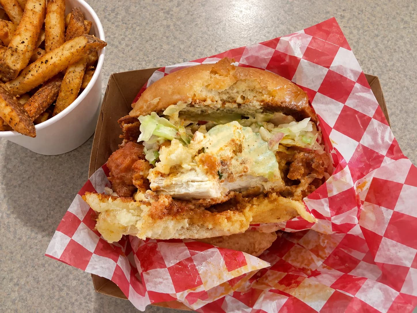 Fried chicken sandwich cut in half on greaseproof paper with a thick craggy-crusted fillet overhanging a soft bun, pickles visible at the cut face