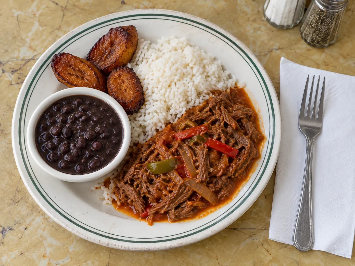 Plate of shredded ropa vieja beef with peppers and onions beside white rice, black beans and fried sweet plantains on a worn diner plate on a laminate counter