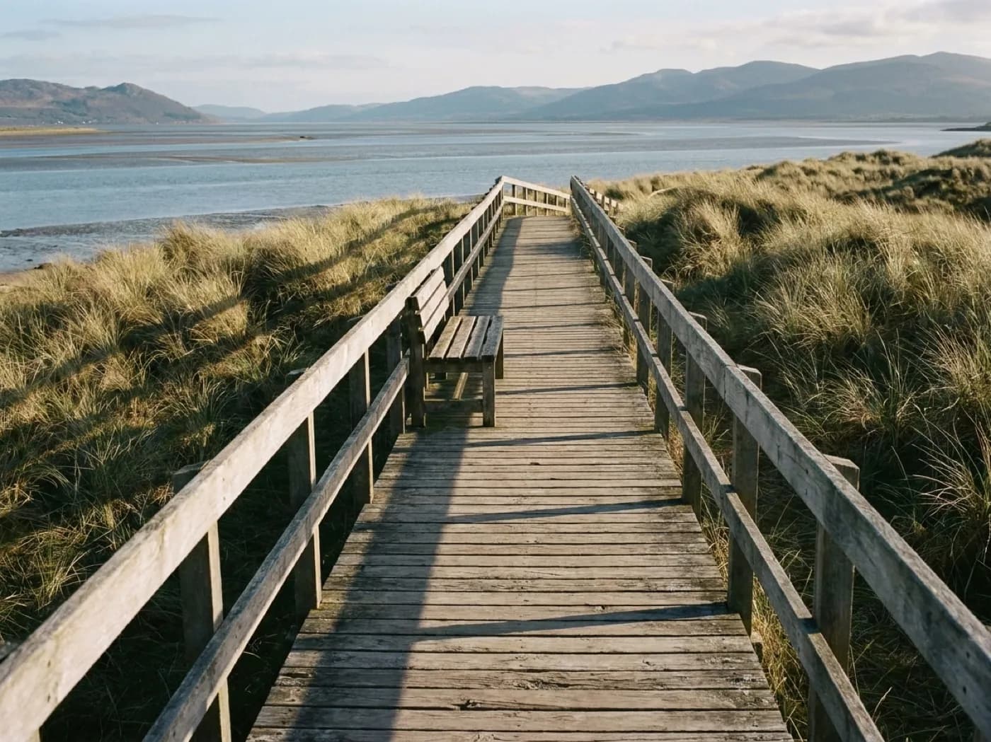 A weathered wooden boardwalk crossing low dune grass toward the water, with a bench and no people