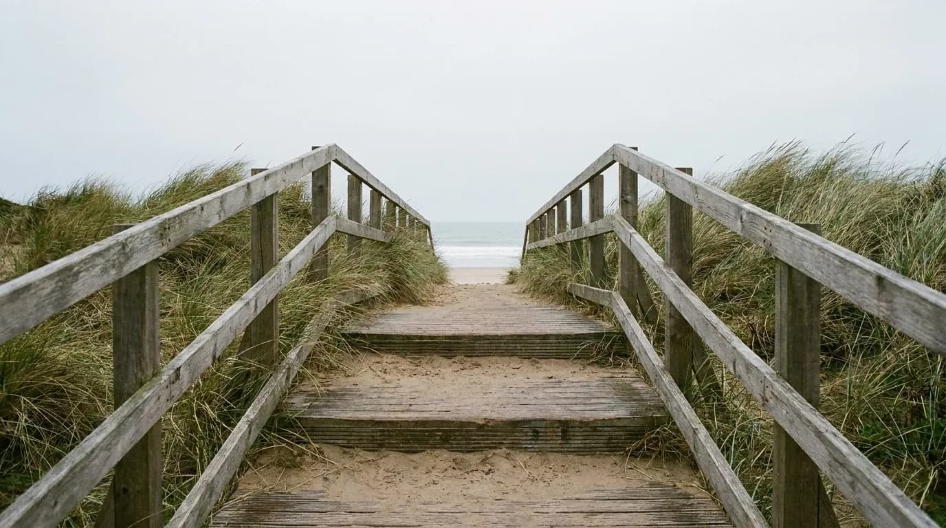 A weathered timber walkway crossing low dunes toward open sand, boards half buried by blown sand and beach grass leaning over both edges