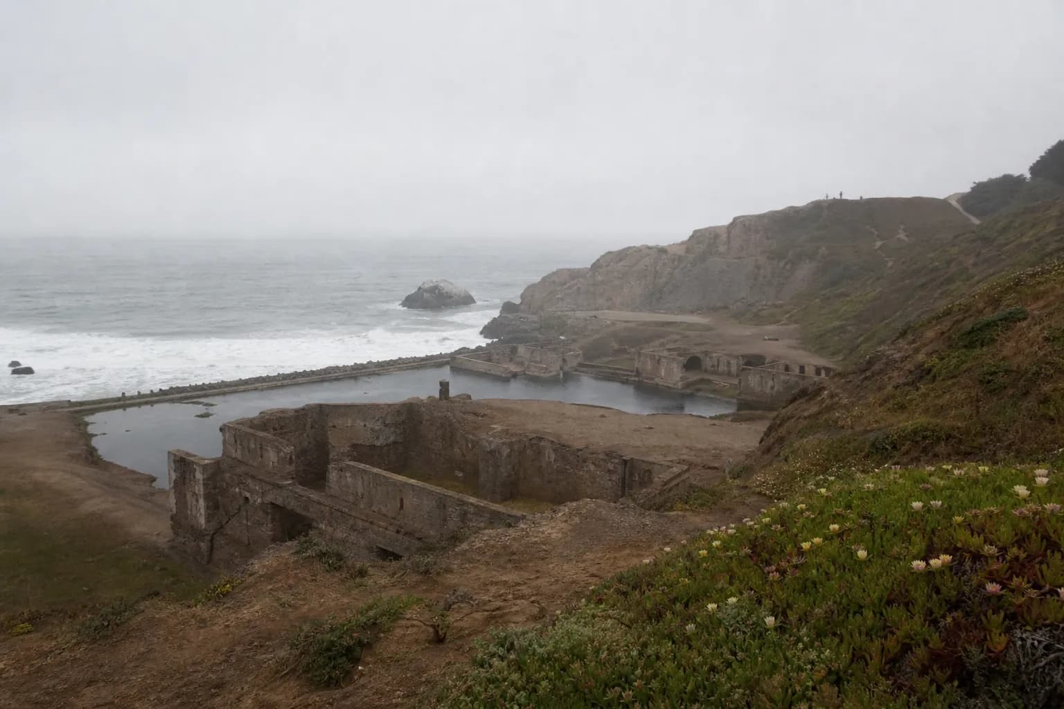 Wide view of Sutro Baths ruins and the trailhead on a gray morning.