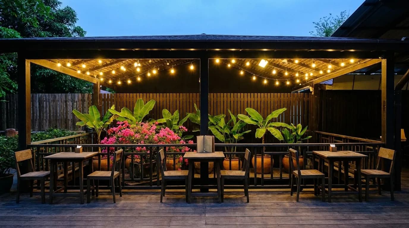 A small restaurant patio behind a low wall on a residential side street at dusk, four tables under string lights with a wooden fence and banana plants behind