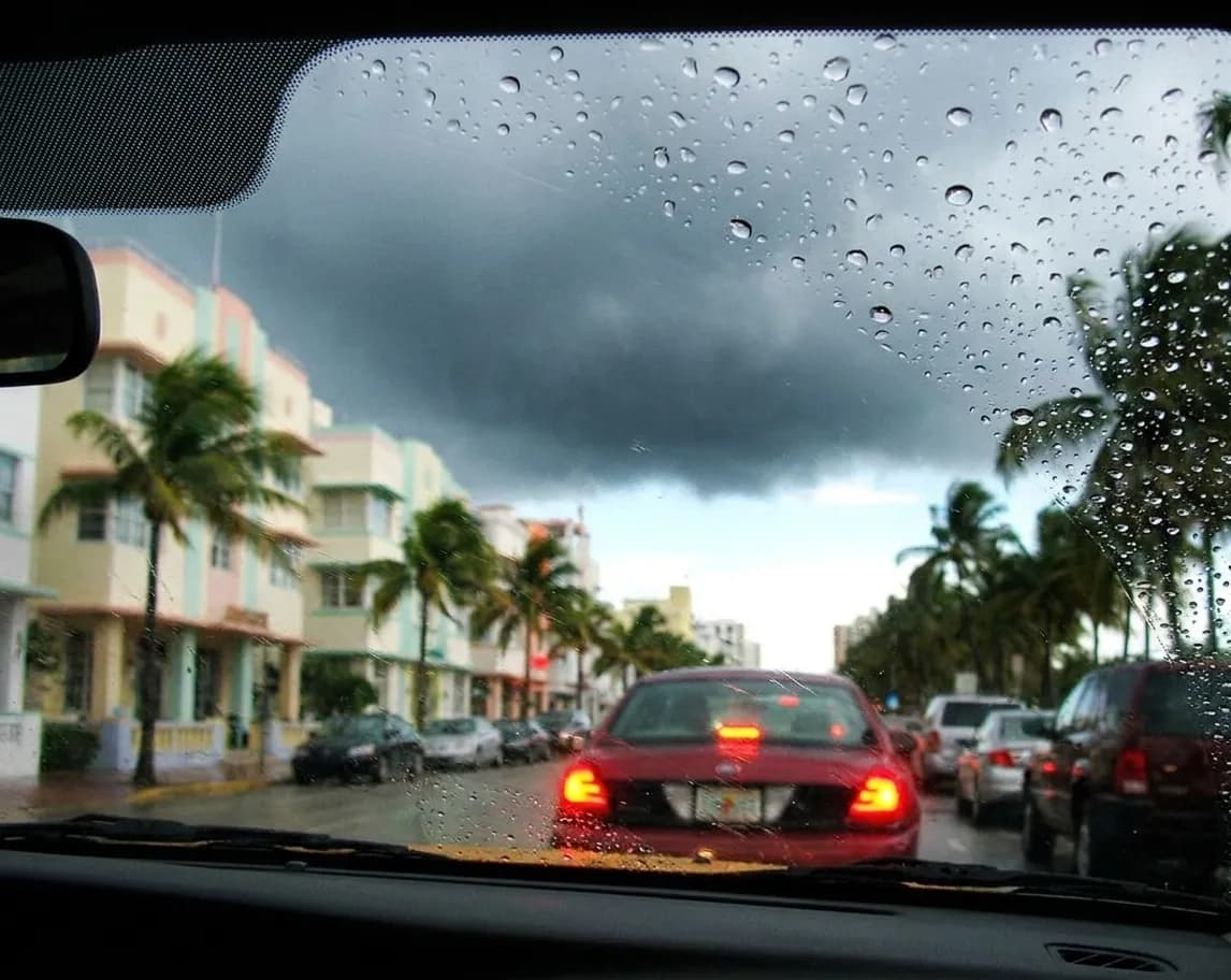 A Miami street seen through a rain-beaded windscreen in an afternoon downpour, blurred pastel buildings and bending palms under storm cloud