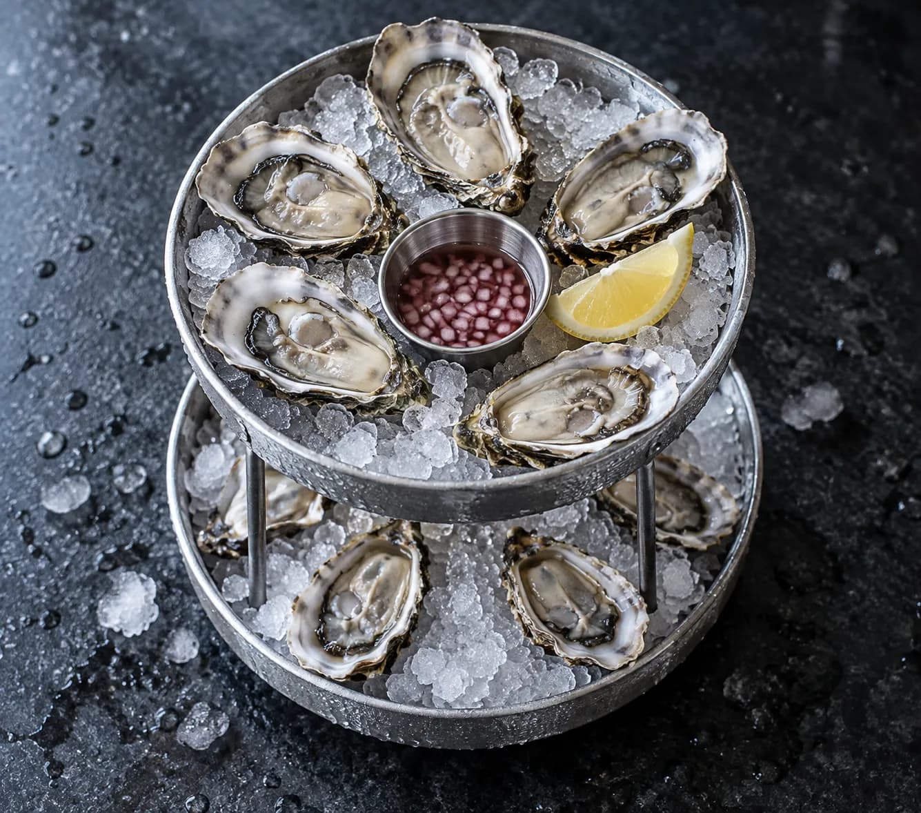 Tiered platter of opened oysters on crushed ice with a lemon wedge and a small pot of mignonette