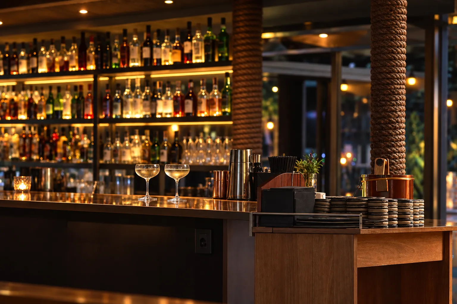 Narrow polished standing bar counter with a back wall of unlabelled shelved bottles under low warm downlights