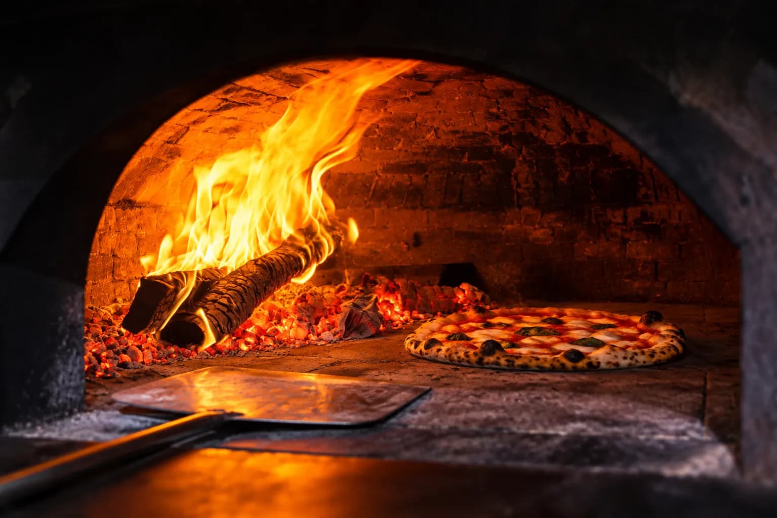 Domed wood-fired oven mid-bake with flames up one side and a blistering pizza on the oven floor