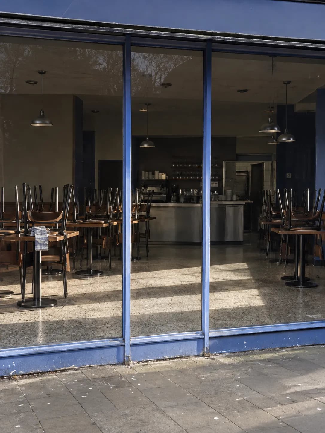 Dark dining room seen through glass with chairs stacked on tables and daylight falling across the floor