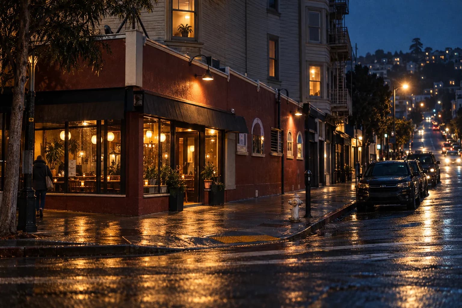 A damp San Francisco commercial street at night, one restaurant front glowing warm among darker shopfronts with hills rising in the distance