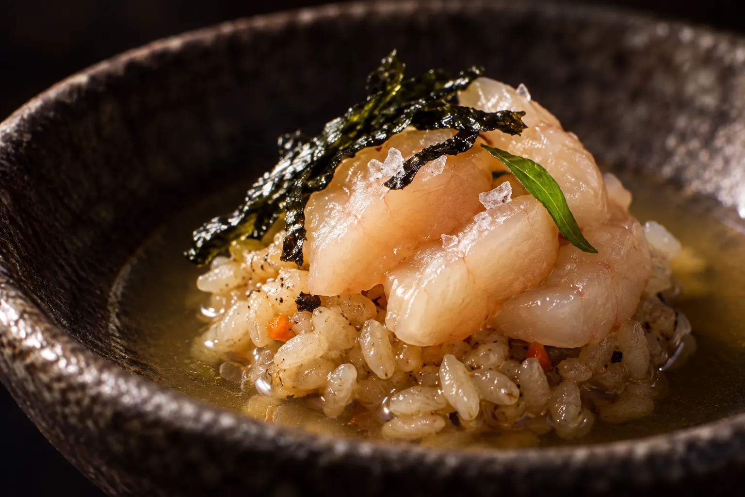 A macro of a single tasting-menu course: three precise slices of raw fish over seasoned rice on a dark hand-thrown ceramic dish in a pool of clear dashi