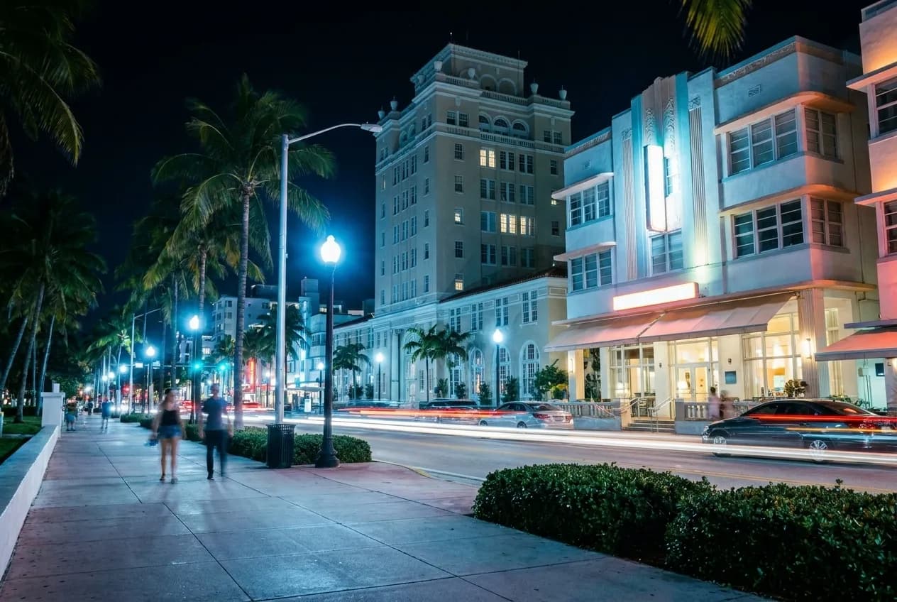 Collins Avenue in South Beach at night with art deco buildings, palm trees, and pedestrians on the sidewalk.