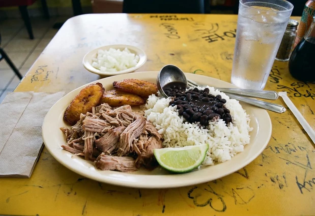 A Cuban plate on a laminate restaurant table with roast pork, white rice, black beans, fried plantain and a lime wedge beside a glass of iced water
