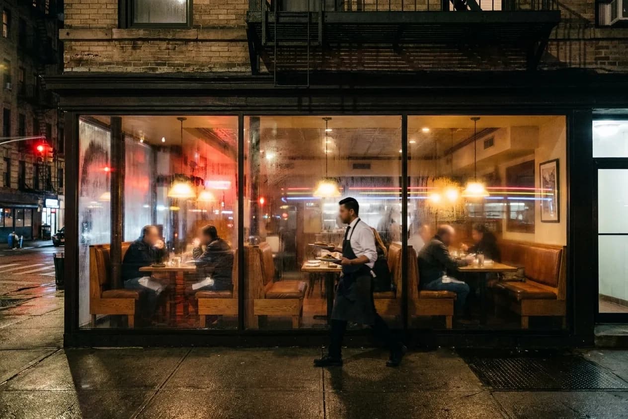 A warm-lit New York restaurant seen through its window near midnight with a few late diners in booths
