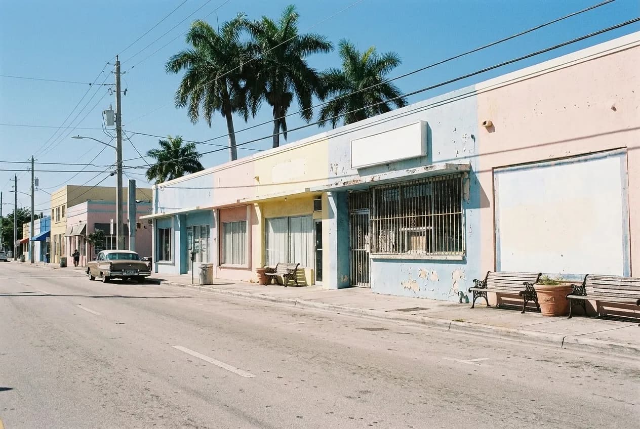 A low-rise commercial block on a wide Miami street in hard midday sun, painted stucco frontages, a barred service window and royal palms breaking the roofline