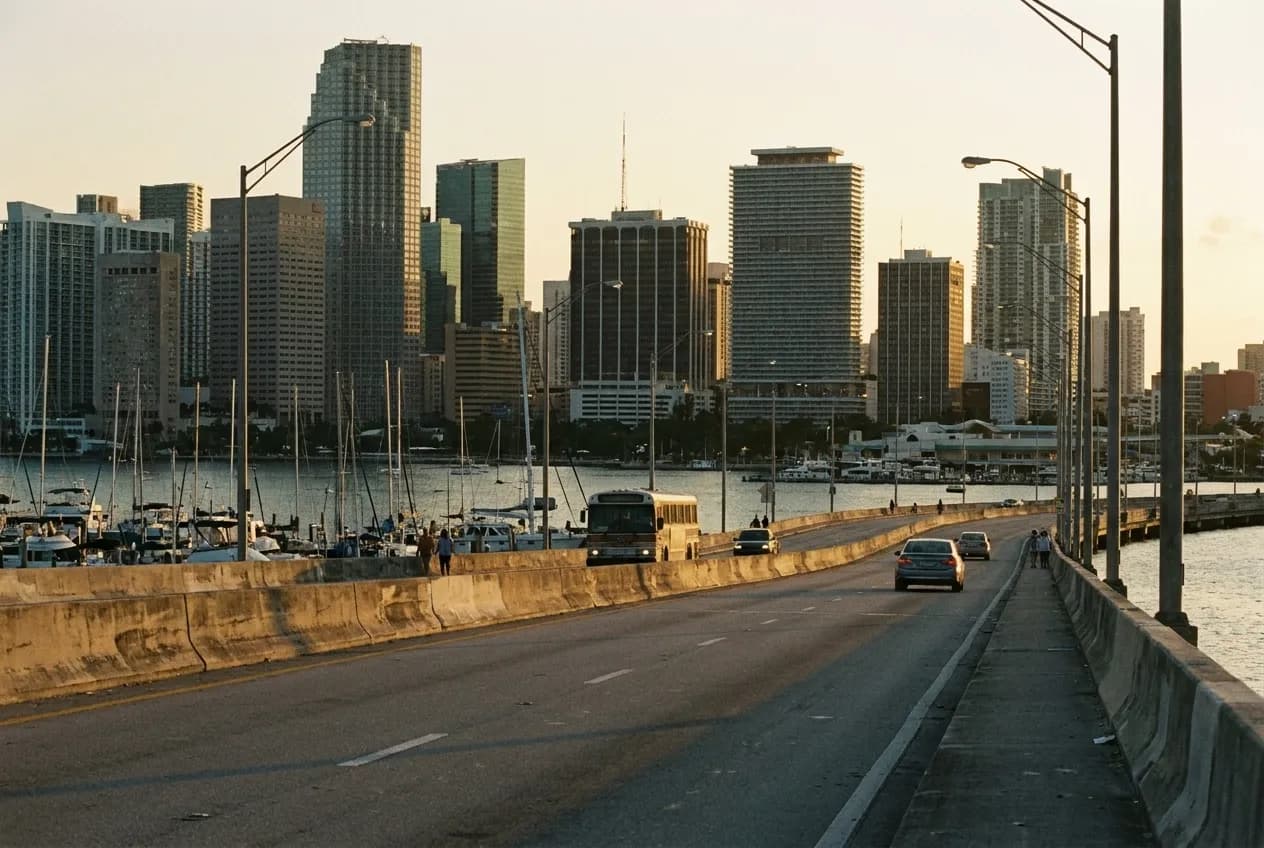 A low causeway crossing Biscayne Bay in late afternoon light, downtown Miami towers stacked on the far side and boat masts in the channel below