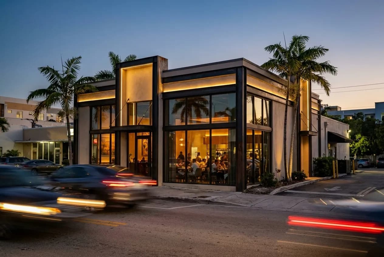 A low-rise stucco building with palm trees and glass storefronts in Miami's Design District at dusk, car headlights moving past on the street.