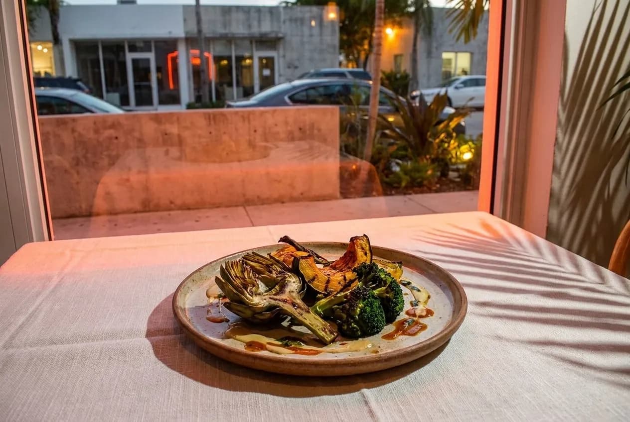 A dinner plate with roasted vegetables and micro-greens on a white linen table, warm evening light through a Miami storefront window behind it.
