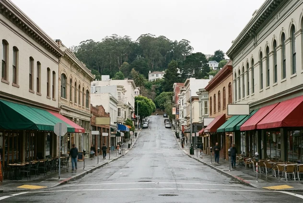 A wide diagonal avenue of low pre-war buildings under flat grey light, awnings over pavement tables and a wooded hill rising at the end of the street
