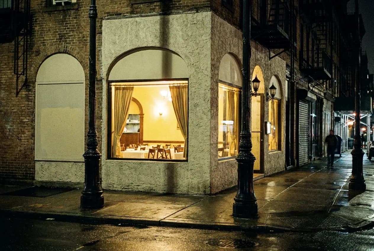 A lit restaurant window on a dark New York street late at night, empty pavement in front