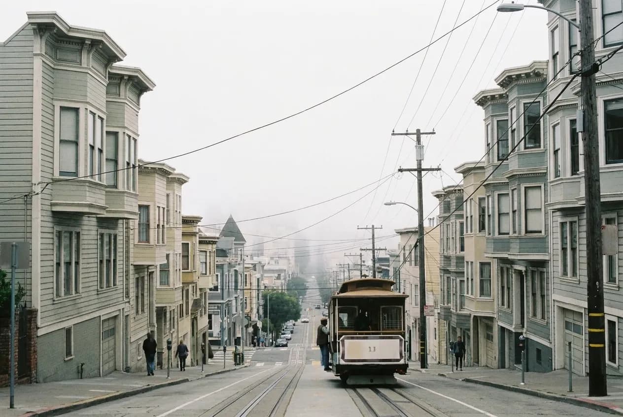 A steeply pitched San Francisco street dropping away toward low fog, painted timber facades stepping down the slope and overhead trolley wires crossing the frame