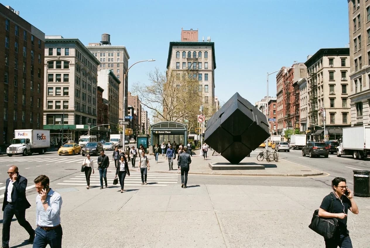 Astor Place intersection at lunchtime with pedestrians crossing toward subway entrance and cube sculpture.