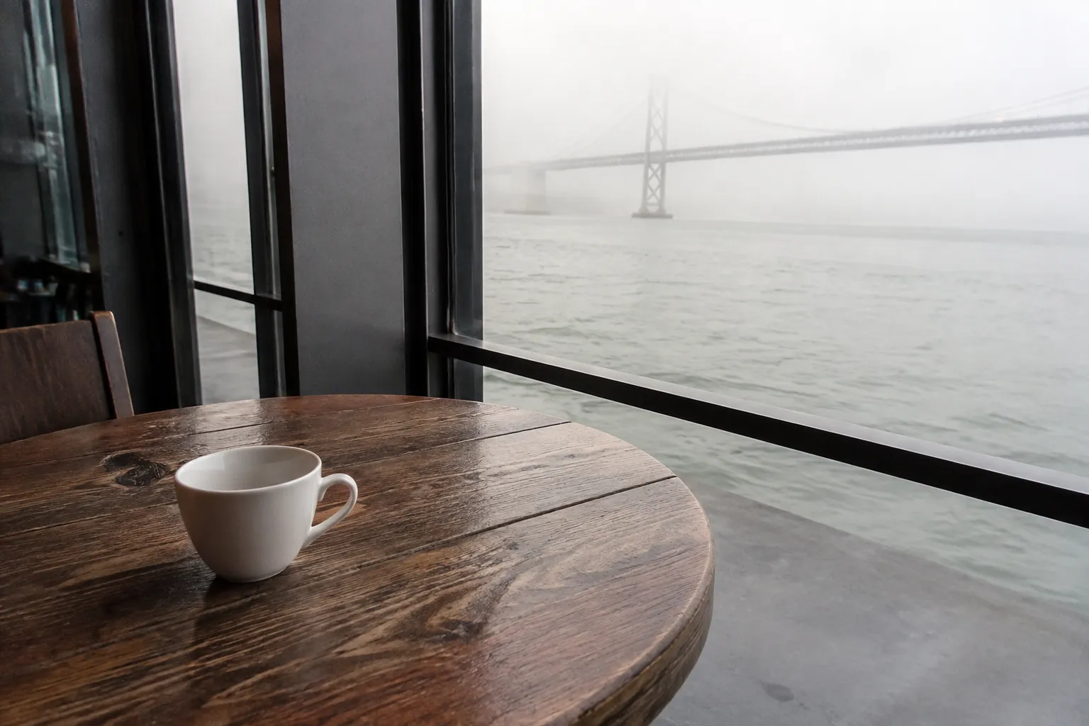 A coffee cup sits on a wooden table near tall windows overlooking the San Francisco waterfront