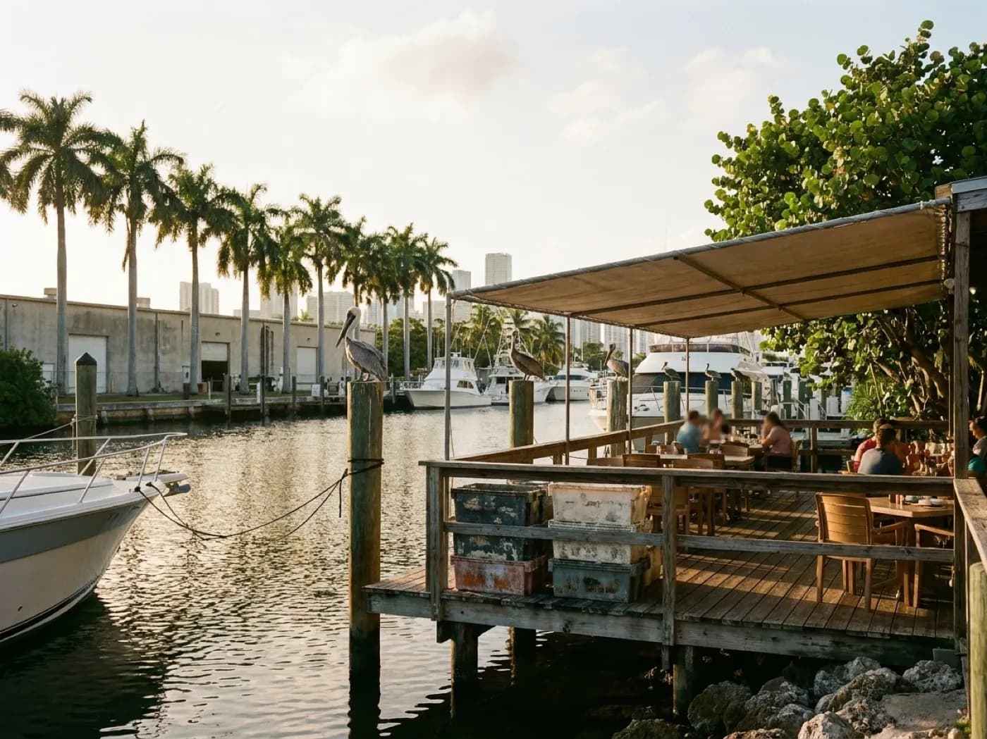 A dockside seafood restaurant on the Miami River at golden hour with wooden tables, moored boats behind and pelicans on the pilings