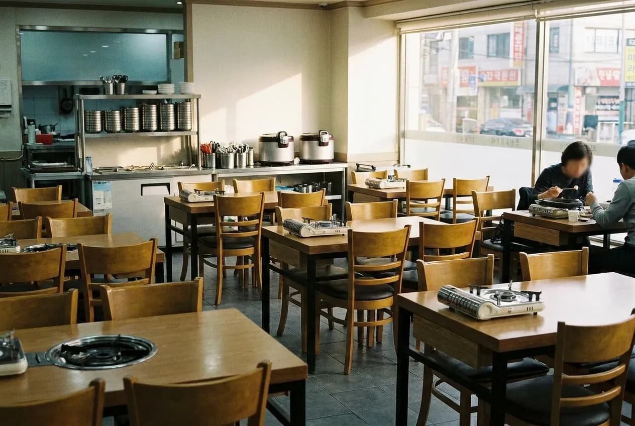 A plain Korean restaurant dining room with burner-set tables and flat daylight through a wide street window