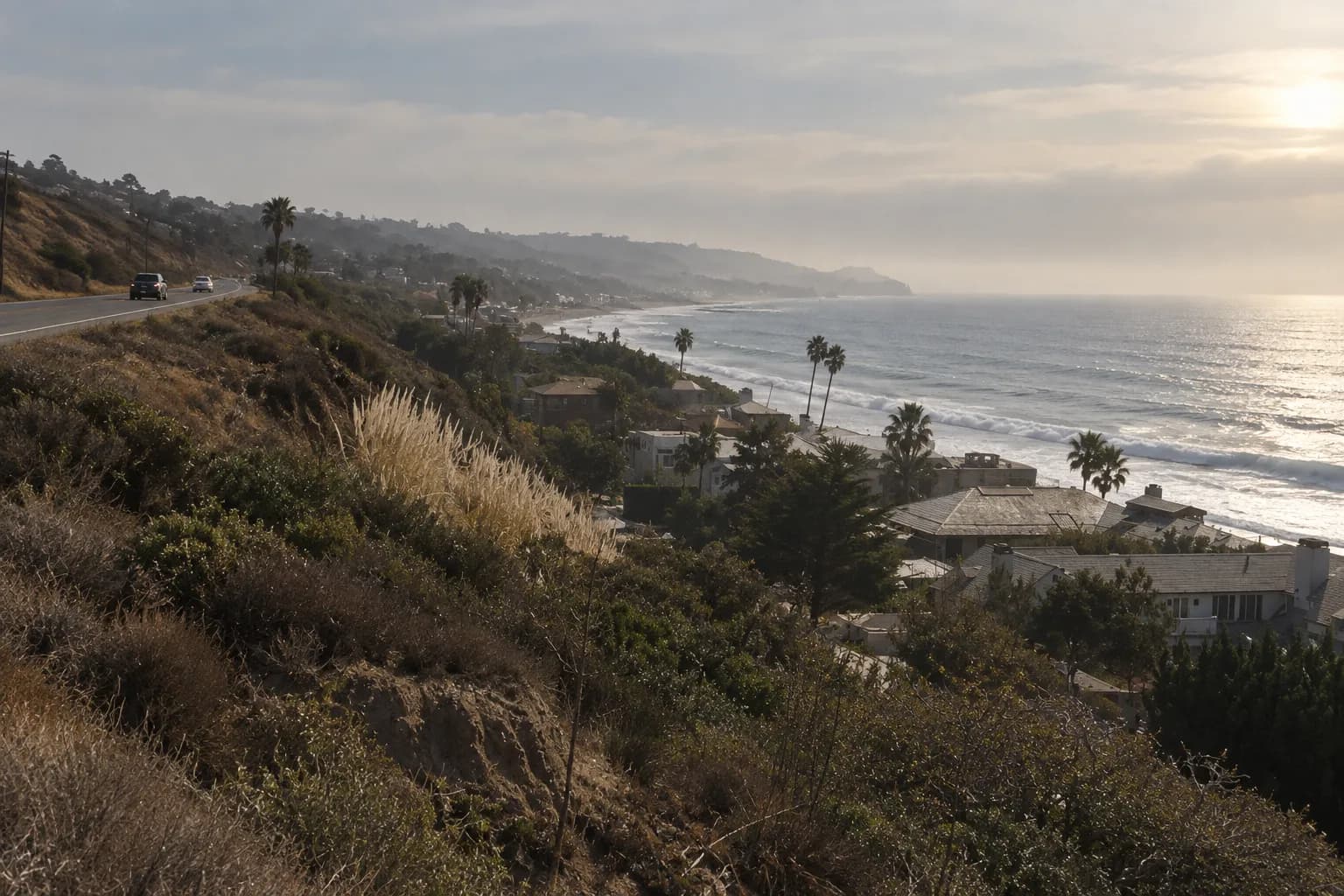 A coast highway in late afternoon with grey-blue ocean on one side, dry brown hills on the other and marine haze softening the horizon