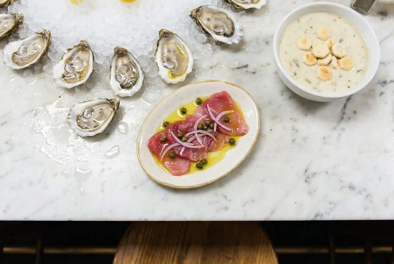 Overhead view of fresh oysters on ice and raw sashimi on a marble counter with a bowl of clam chowder.