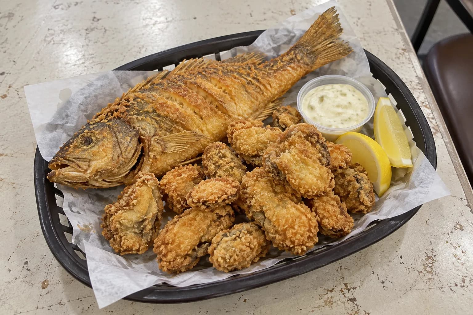 A paper-lined plastic basket holding a whole fried fish and fried shellfish pieces with lemon wedges and a pot of pale sauce on formica