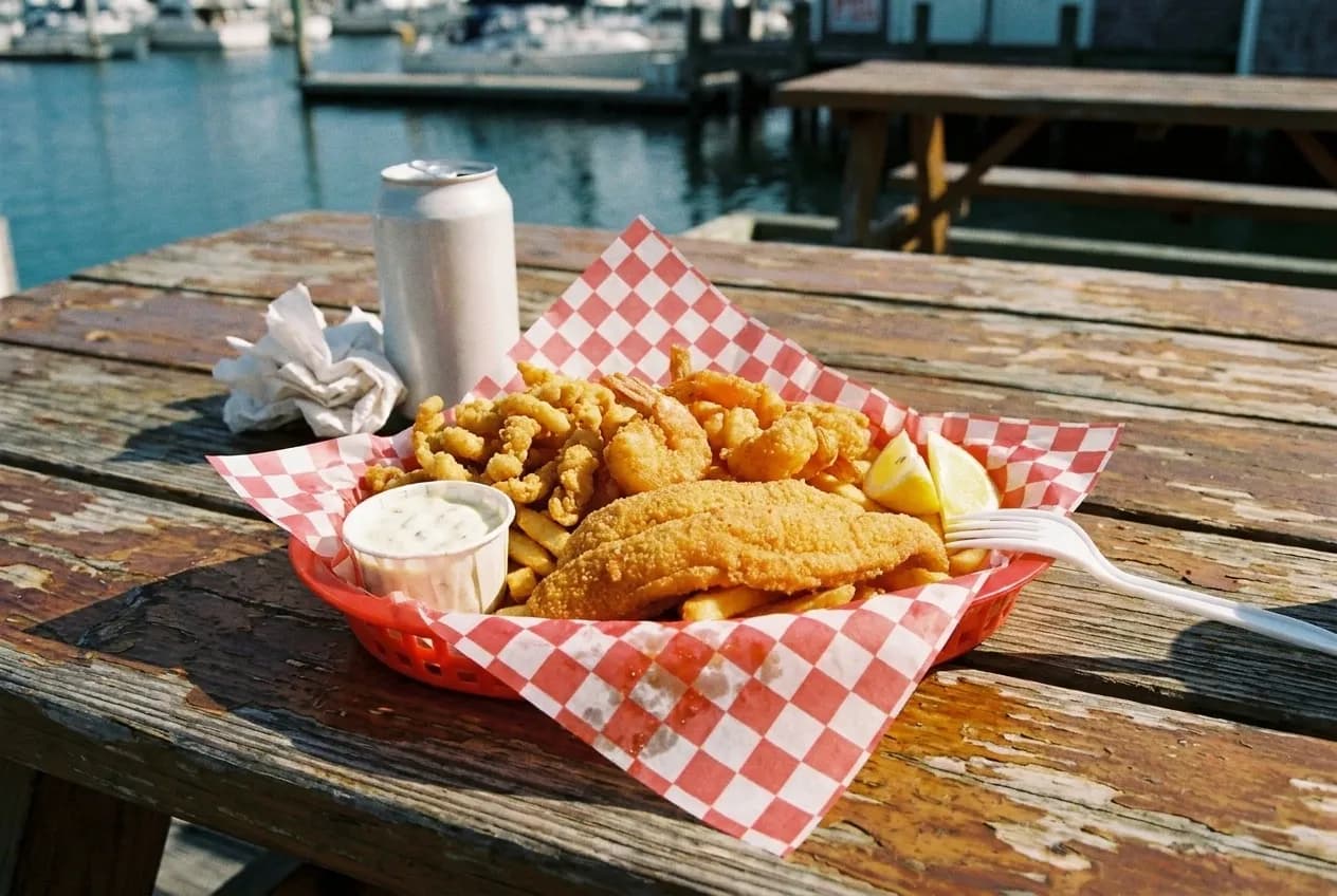 Fried seafood basket with fish, shrimp and clam strips in a red plastic basket on a weathered picnic table beside City Island's moored boats