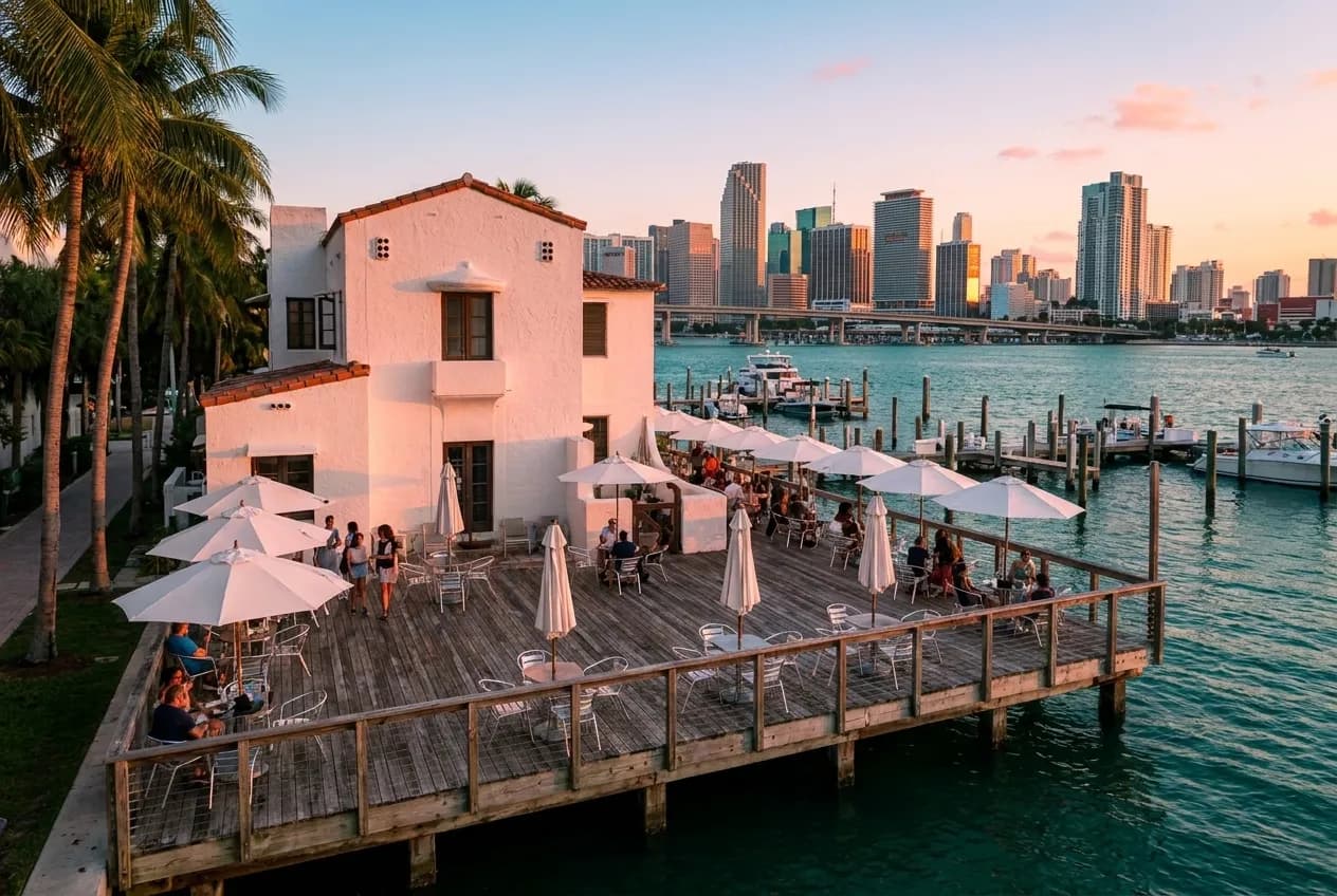 Wide wooden deck with white umbrellas and metal chairs extending over Biscayne Bay at golden hour, Downtown Miami skyline rising across teal water under pink-orange light, palms framing the left edge.